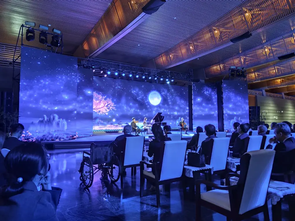 Gala cultural con músicos en escenario ante una gran pantalla LED que proyecta un paisaje de luna y cerezo, frente al público.